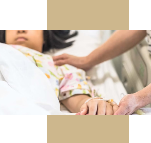 Child lying in a hospital bed wearing a patterned gown, with an IV line in their hand. An adult beside the bed gently holds the child’s hand in a gesture of comfort and care.