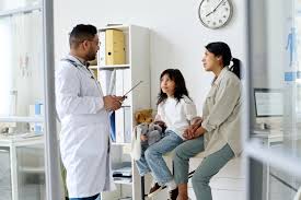 A doctor in a white coat reviewing a clipboard while seated across from a parent and child in a bright clinic room; the child sits on the exam table and the parent stands nearby, with shelves and medical equipment visible in the background.
