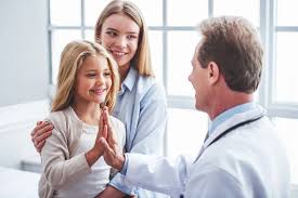 A smiling young girl gives a high five to a doctor in a white coat while her mother stands behind her, smiling. They are in a bright clinic room with large windows and natural light.