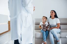 A woman and her young daughter sit together on a beige couch in a bright, modern clinic. The child holds a teddy bear and looks up at a doctor standing nearby, while the mother listens attentively. The setting is clean and minimalist with white walls and natural light.